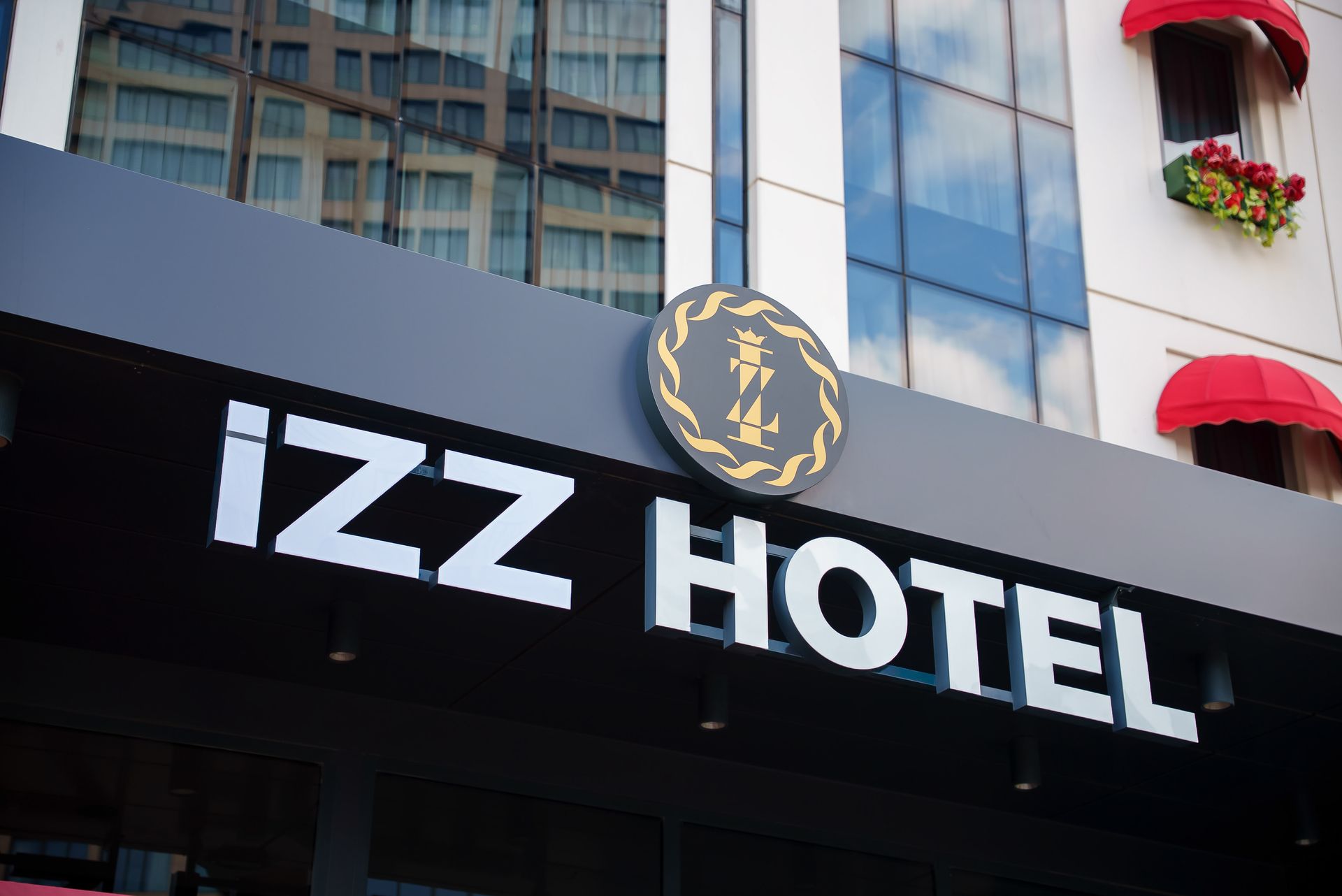 Modern bir bina cephesinde, yansıtıcı cam pencereleri ve kırmızı tenteleriyle IZZ Otel tabelası.