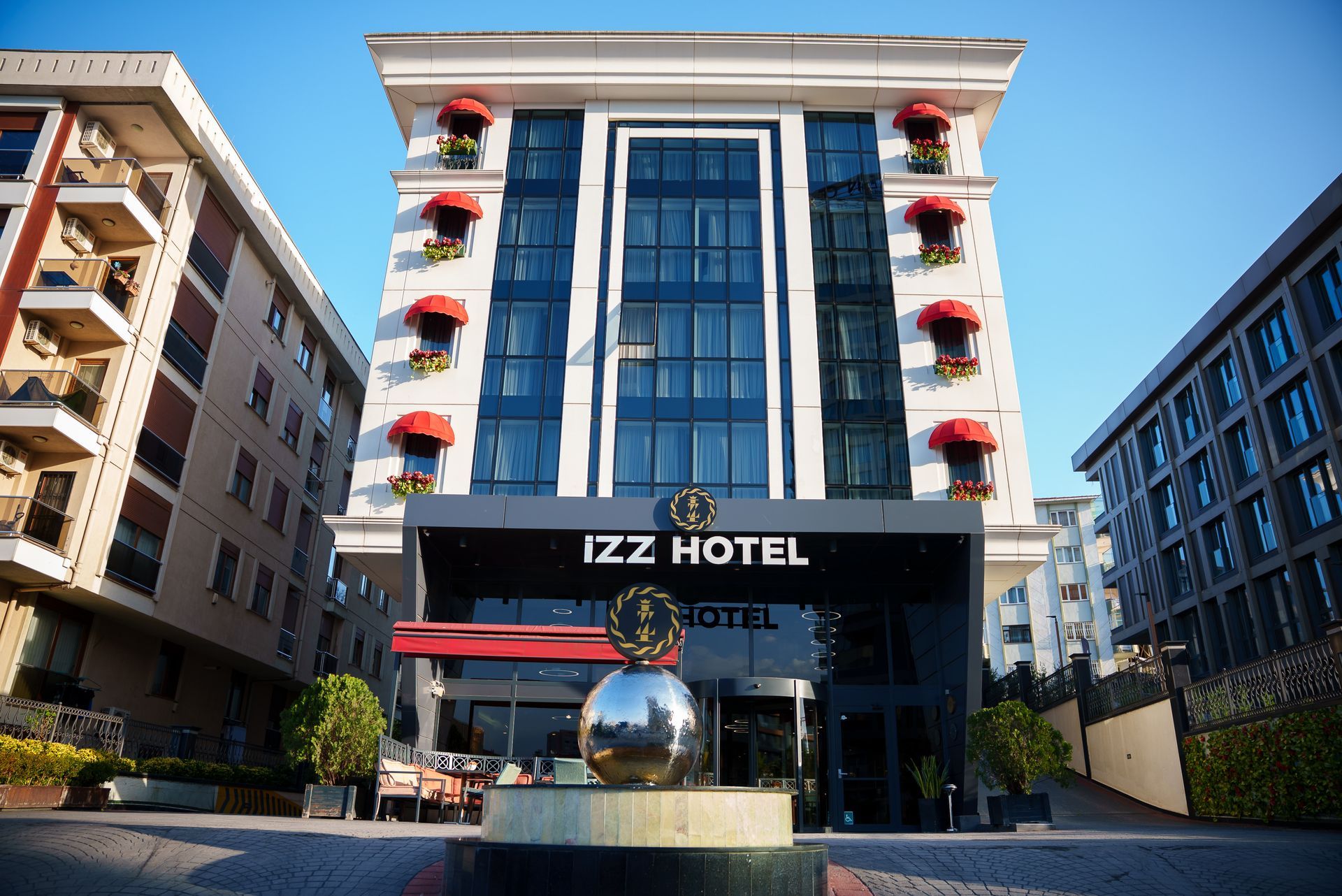 Beyaz cephesi, kırmızı balkon çiçekleri ve mavi cam girişiyle Izz Otel binası, berrak bir gökyüzü altında.