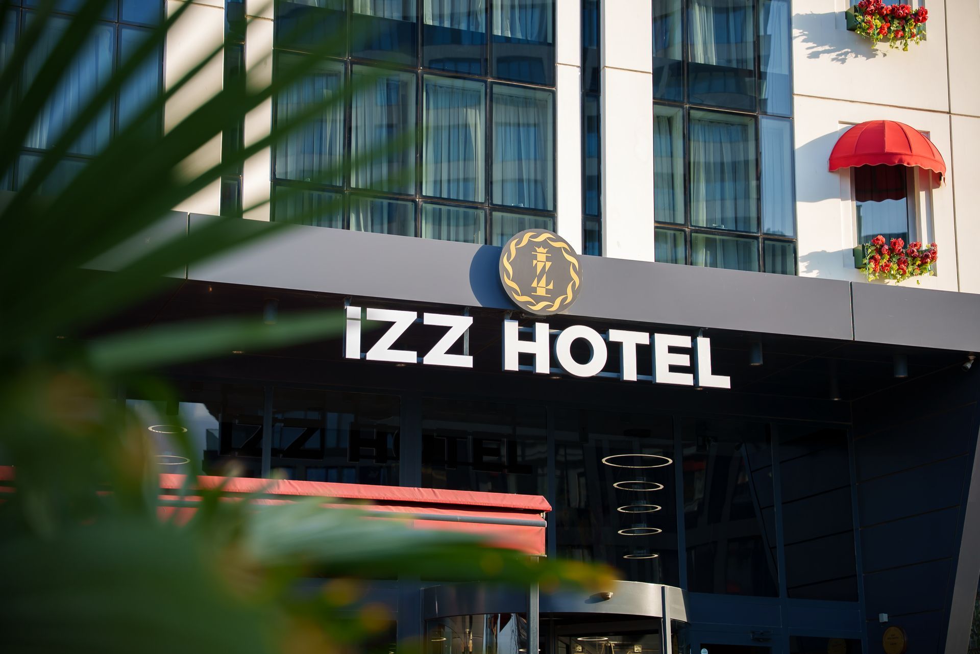 IZZ Oteli'nin girişi, siyah tabelası, cam cephesi ve ön plandaki yaprakların arkasında kırmızı tenteleriyle dikkat çekiyor.