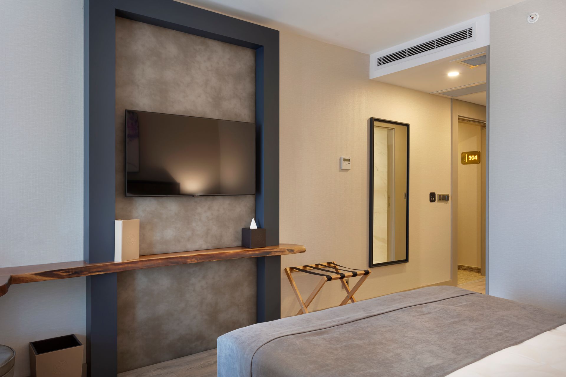 Duvara monte televizyon, ahşap masa, ayna ve yatağın köşesinin görülebildiği modern otel odası.