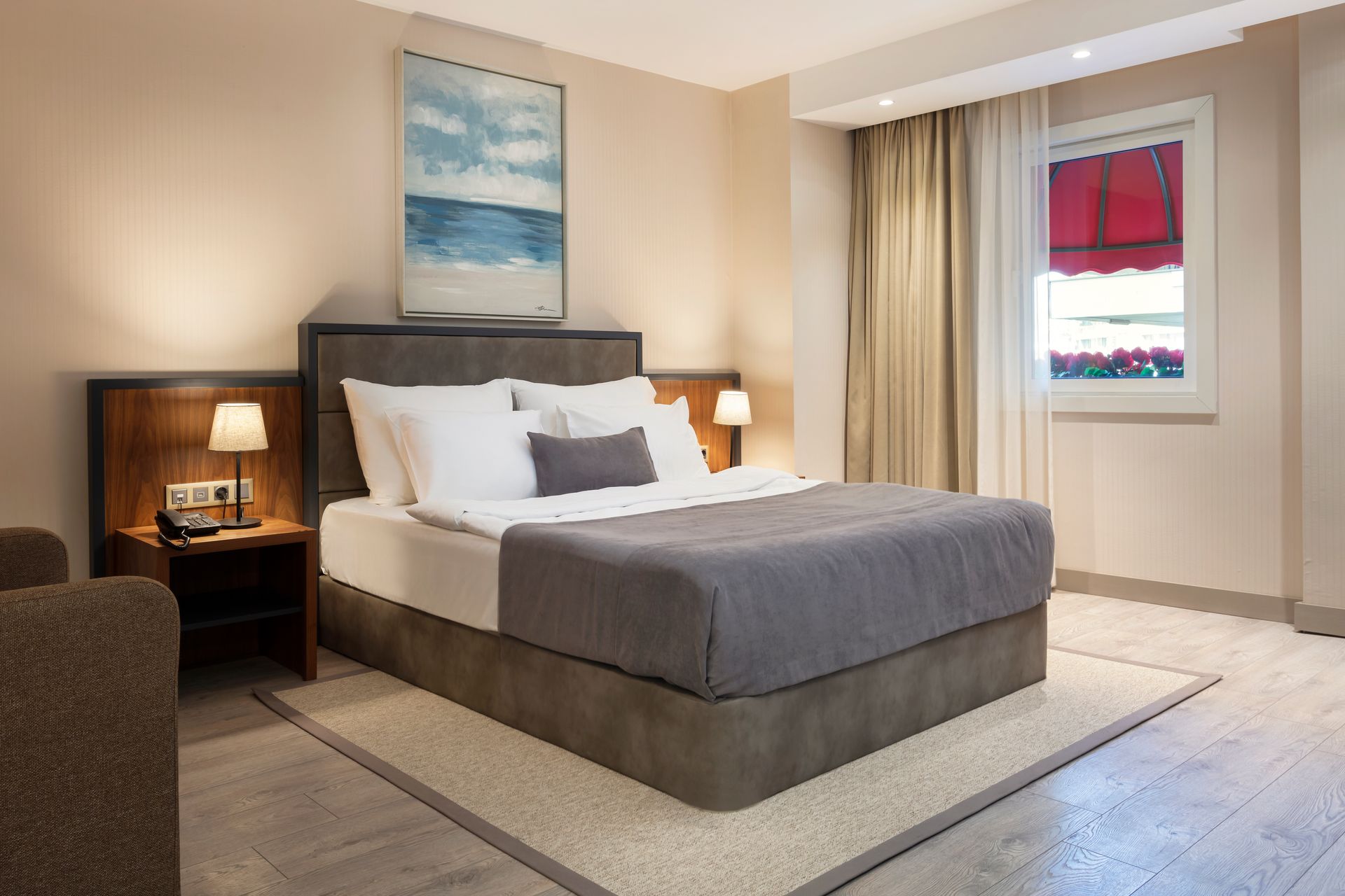 Çift kişilik yatak, komodin lambaları, bej dekorasyon ve kırmızı jaluzili pencereye sahip modern otel odası.
