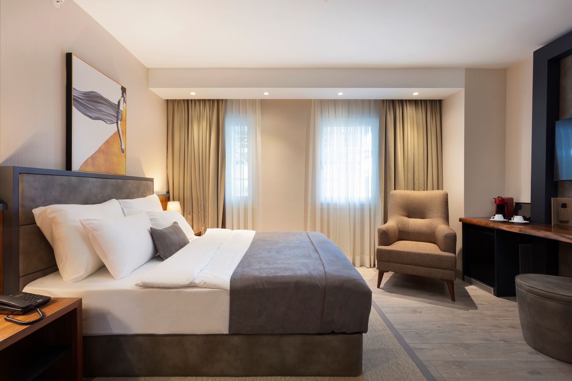 Pencere kenarında yatak, koltuk, çalışma masası ve yumuşak, nötr tonlarda dekore edilmiş modern otel odası.
