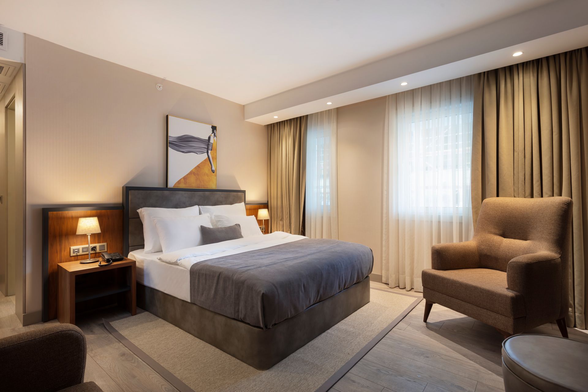 Düzgünce hazırlanmış yatak, başucu lambası, koltuk ve büyük perdeli pencerelere sahip modern otel odası.
