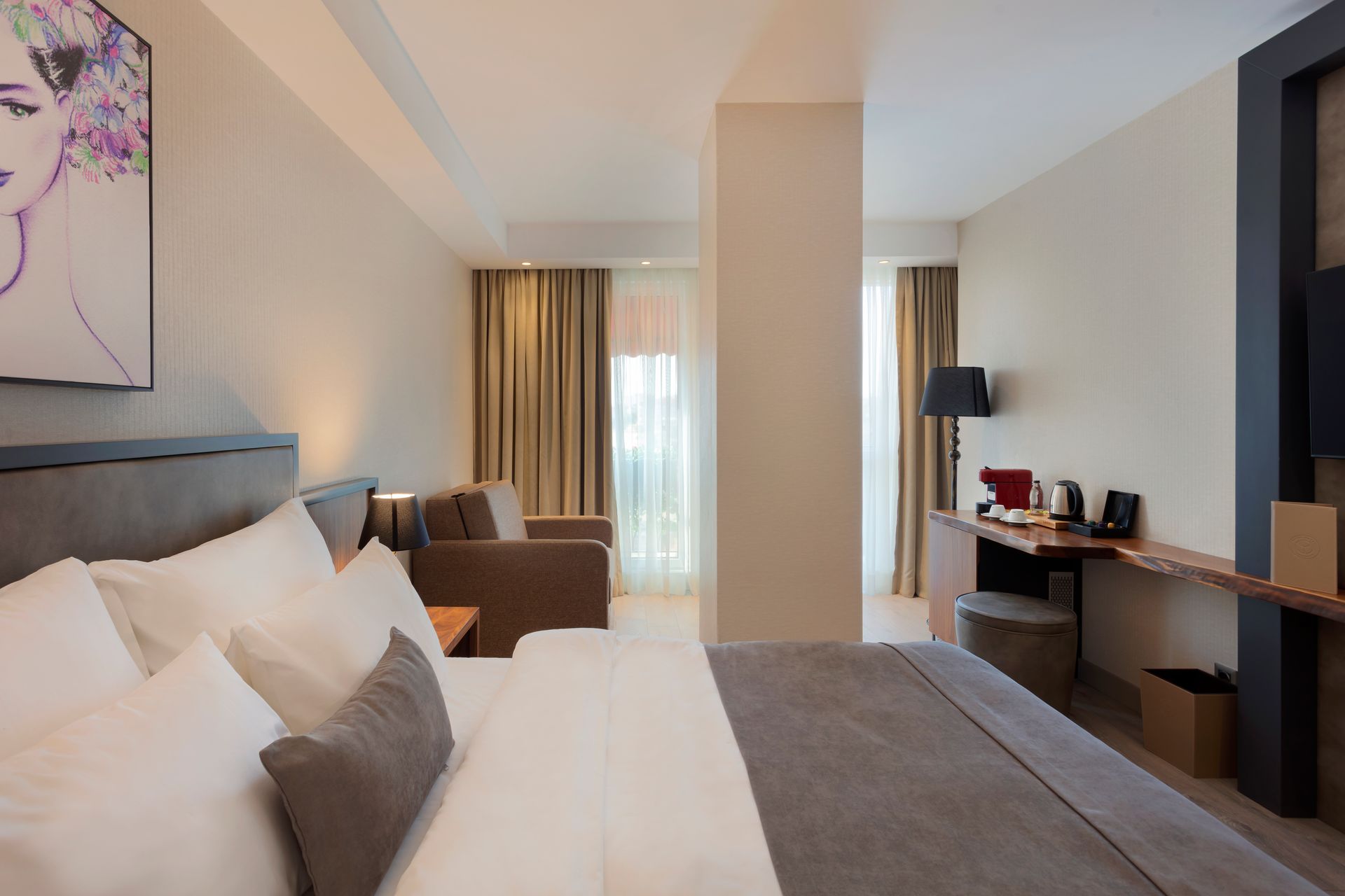Pencere kenarında yatak, kanepe, çalışma masası ve büyük perdeler bulunan modern otel odası.