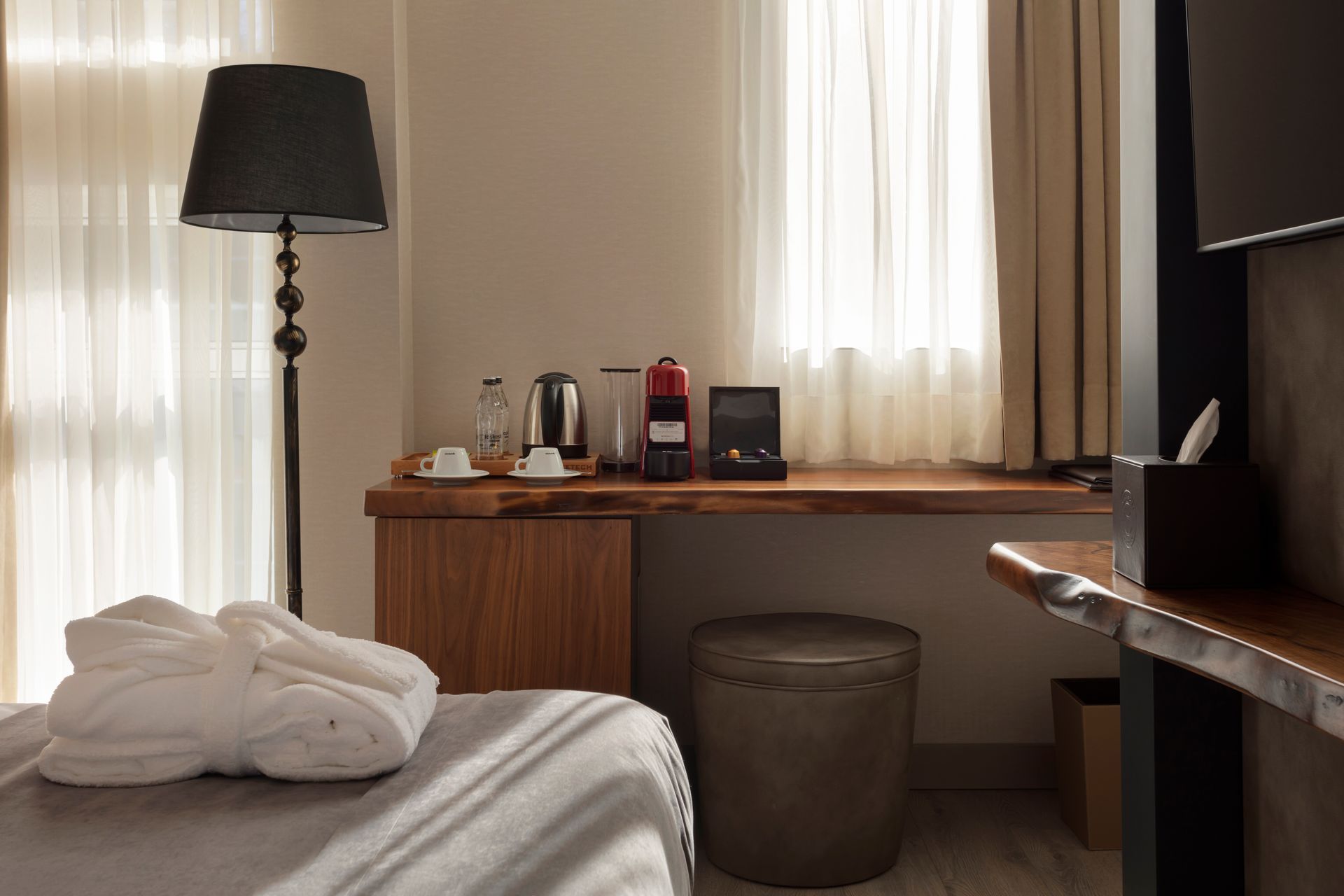 Otel odasında yatak, lamba, çalışma masası ve pencere bulunmakta olup, yatağın üzerinde havlular özenle düzenlenmiştir.