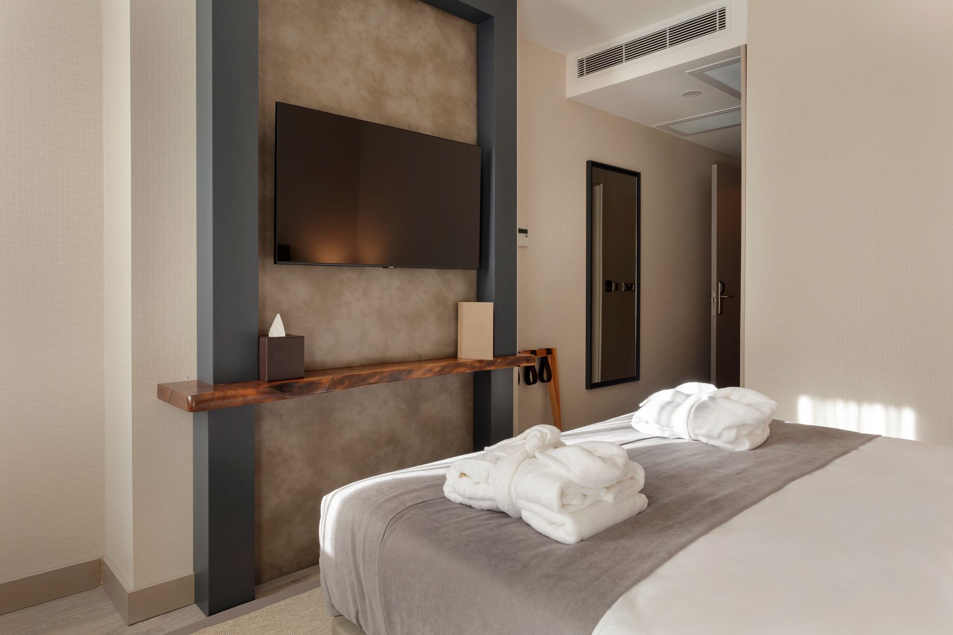 Duvara monte televizyon, ahşap raf ve katlanmış havlularla donatılmış yatak bulunan modern otel odası.