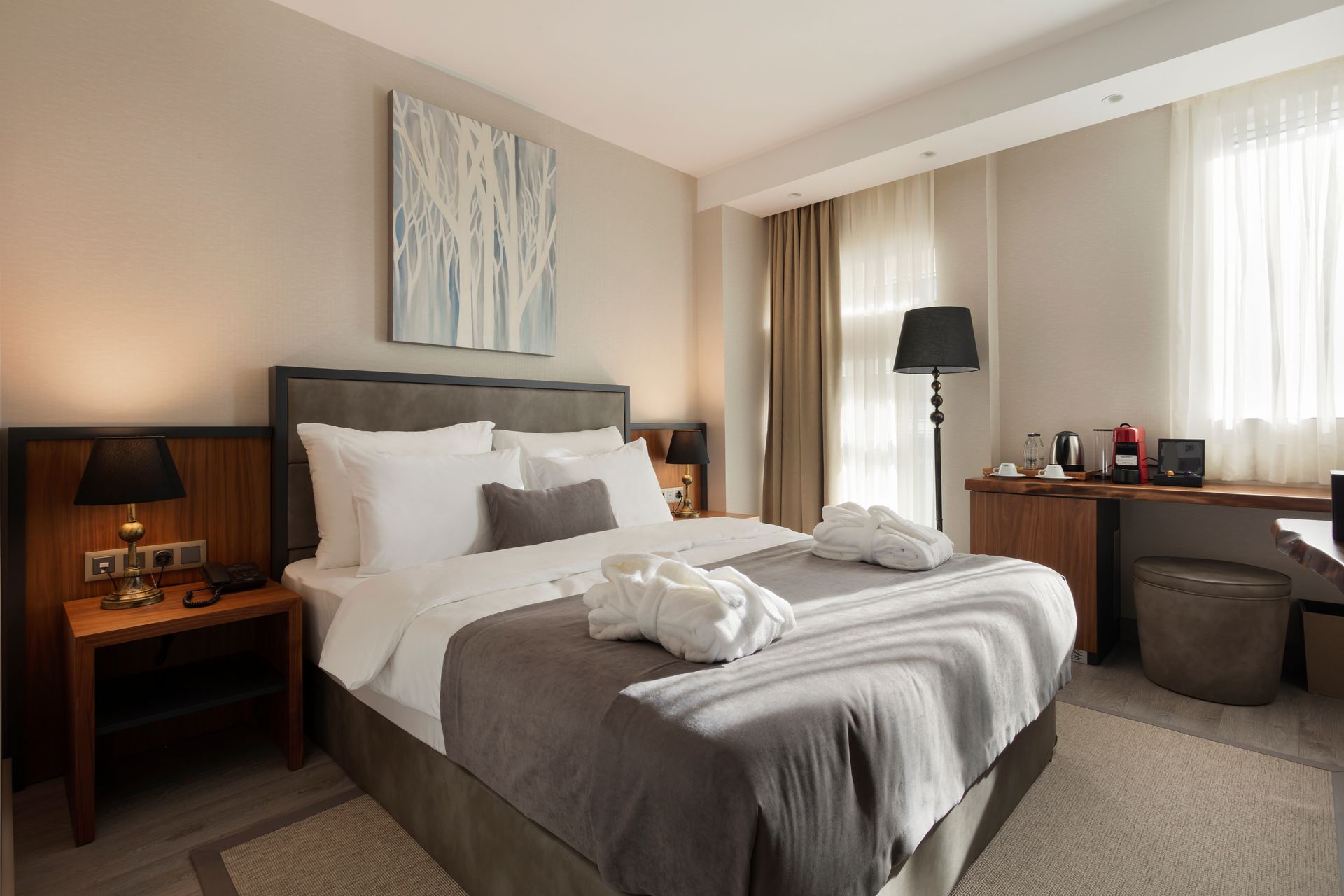 Aydınlık otel odası, özenle hazırlanmış yatak, komodinler, çalışma masası ve perdeli büyük pencerelere sahiptir.