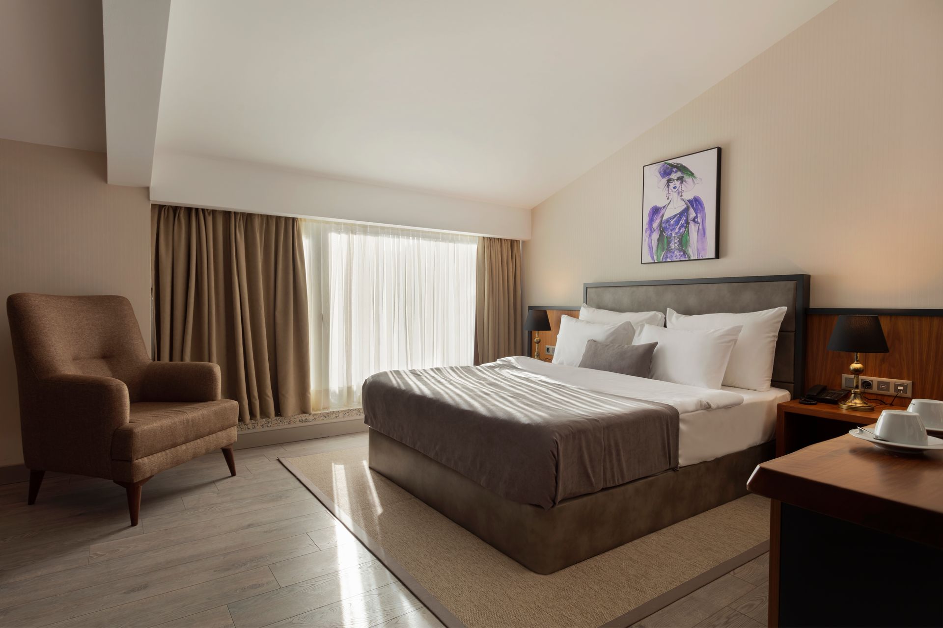 Çift kişilik büyük yatak, koltuk, çalışma masası ve perdeli büyük pencereye sahip modern otel odası.