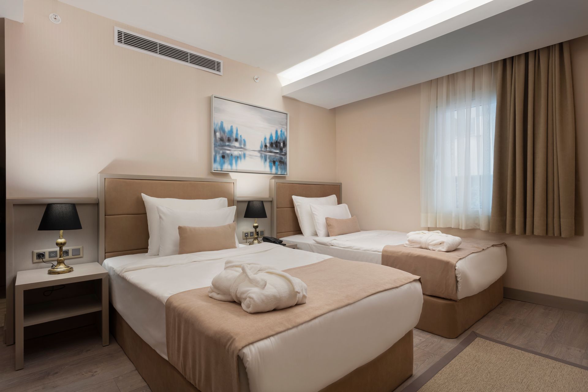 Penceresi ve perdeleri olan modern bir otel odasında, özenle hazırlanmış iki adet bej renkli tek kişilik yatak.
