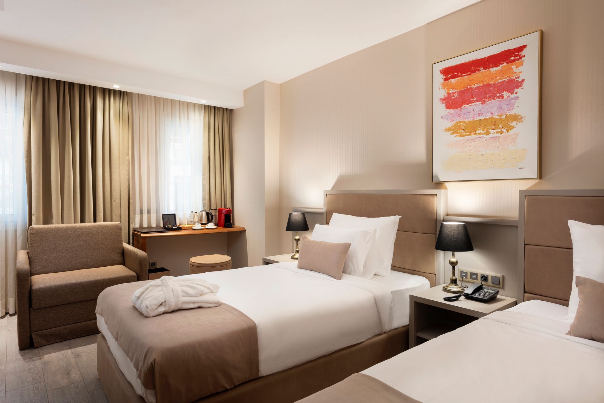 İki yataklı, bej renkli dekorasyonlu, çalışma masası ve sandalyeli, soyut duvar sanatıyla süslenmiş modern otel odası.