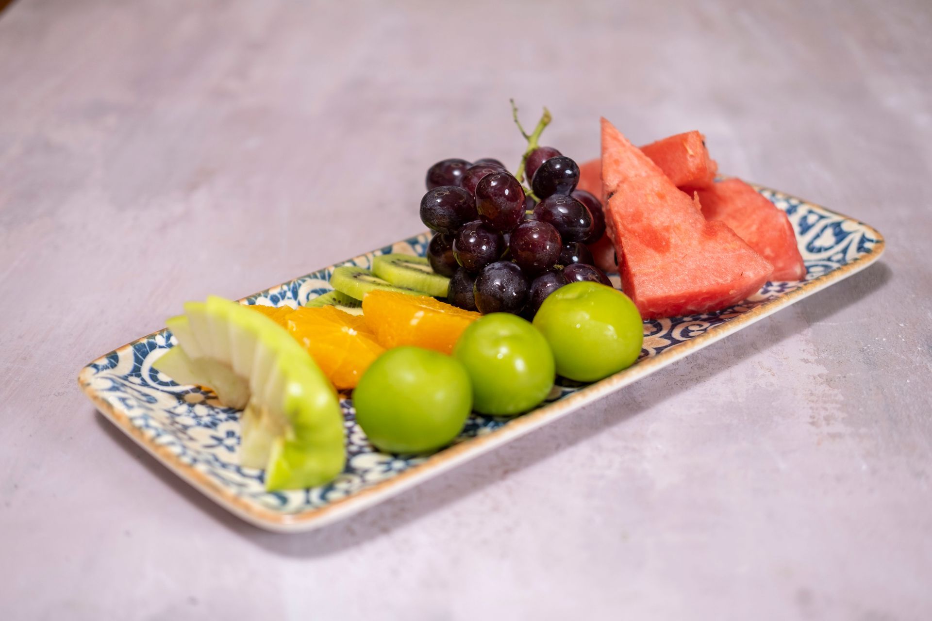 Dikdörtgen bir tabakta çeşitli taze meyveler: üzüm, karpuz, misket limonu ve bir dilim limon.