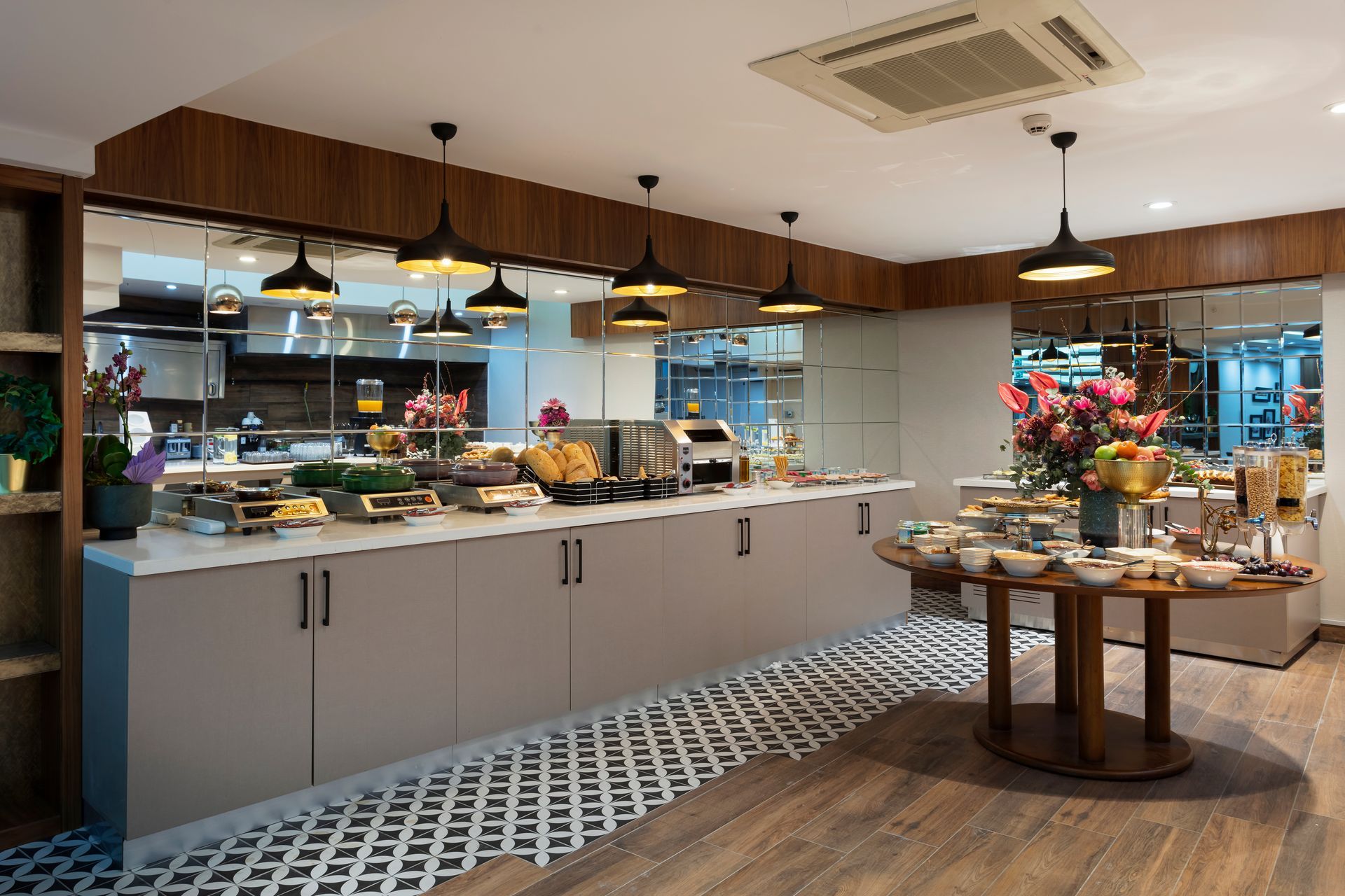 Aydınlık bir kafe iç mekanında, pastalar, çiçekler ve sarkıt lambalarla süslenmiş modern büfe tezgahı.