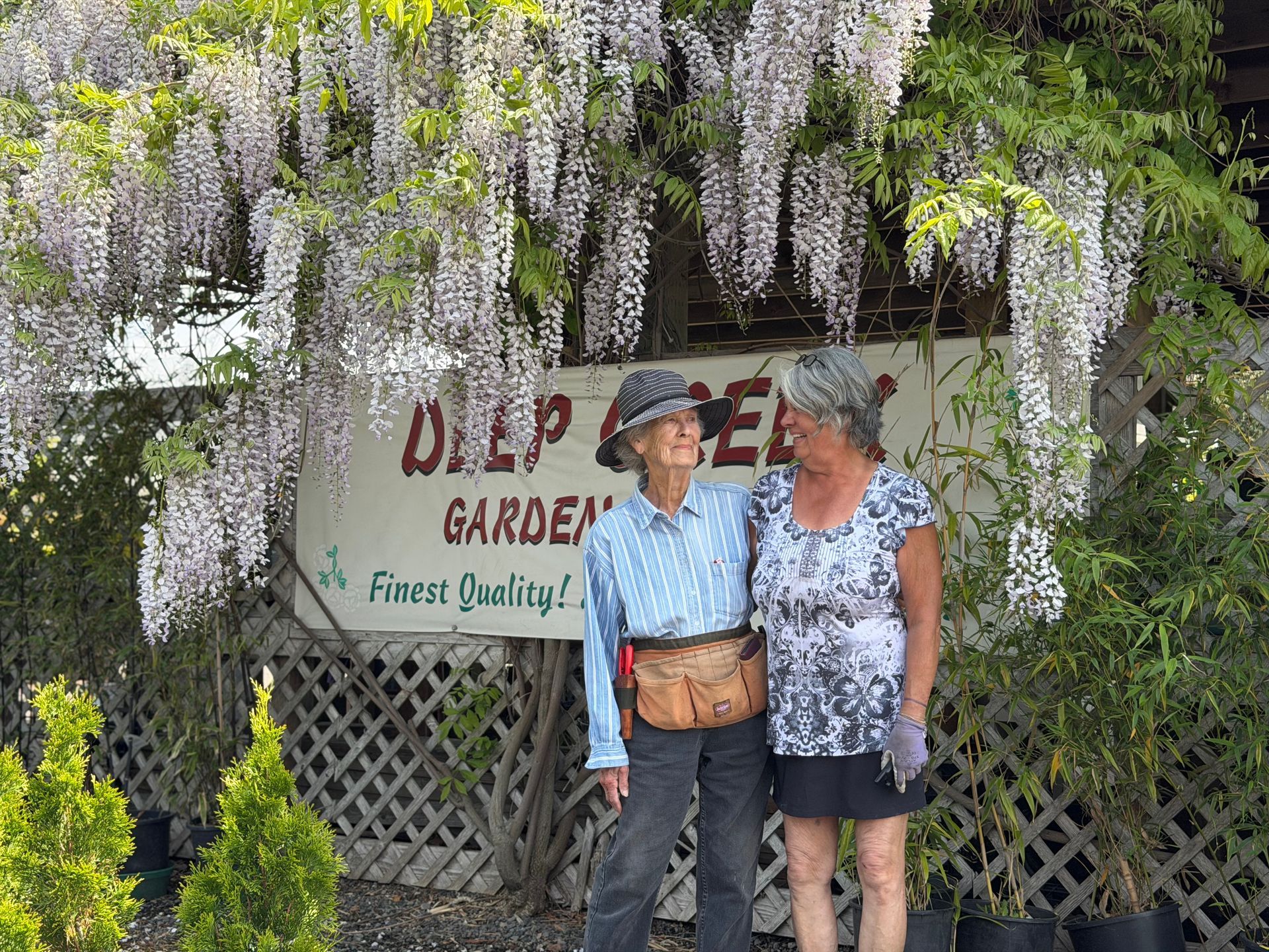 Two people standing under hanging purple flowers in front of a garden sign, smiling at each other.