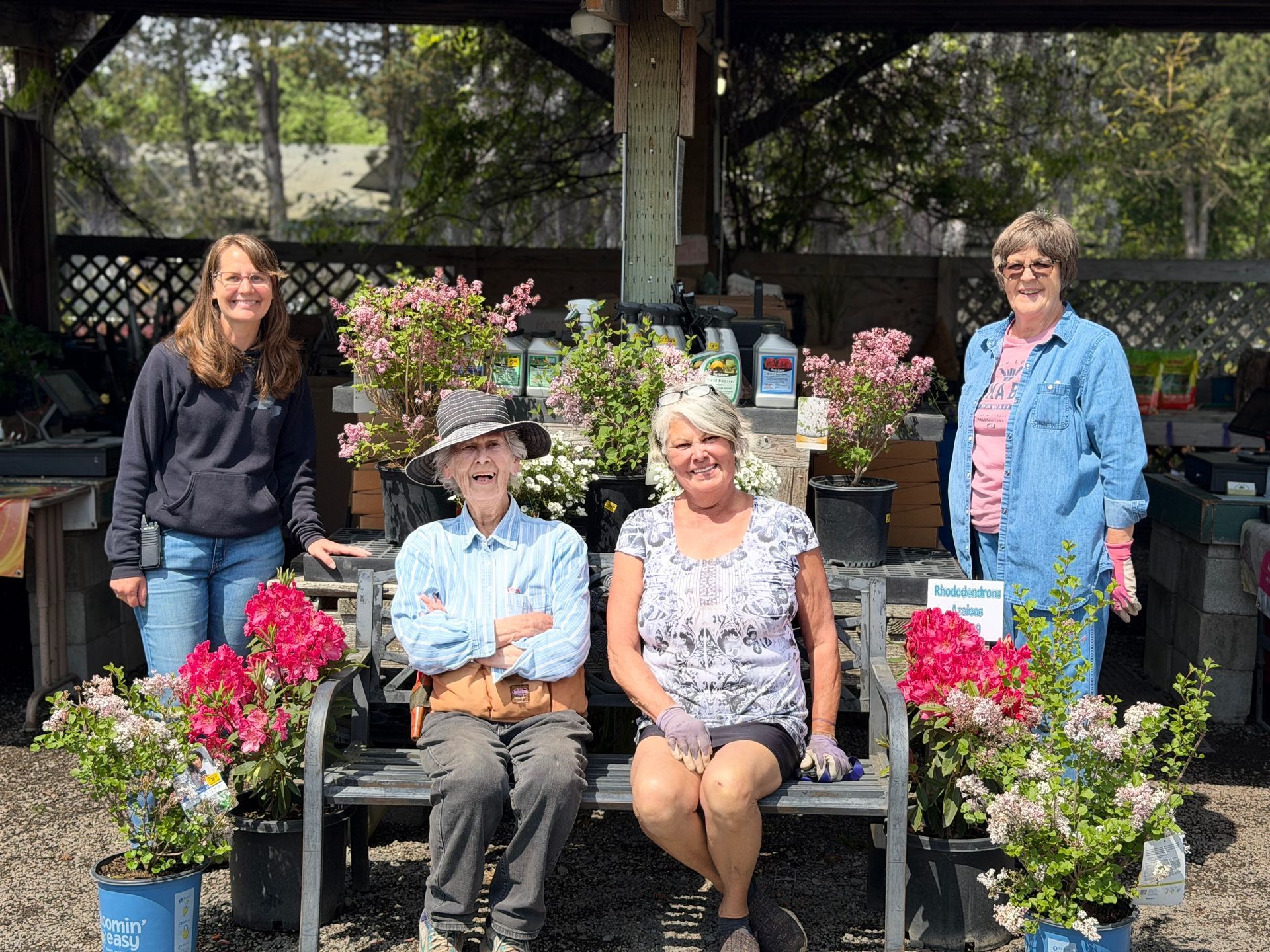 Five people posing among potted flowers in an outdoor garden center, with black benches and trees behind them