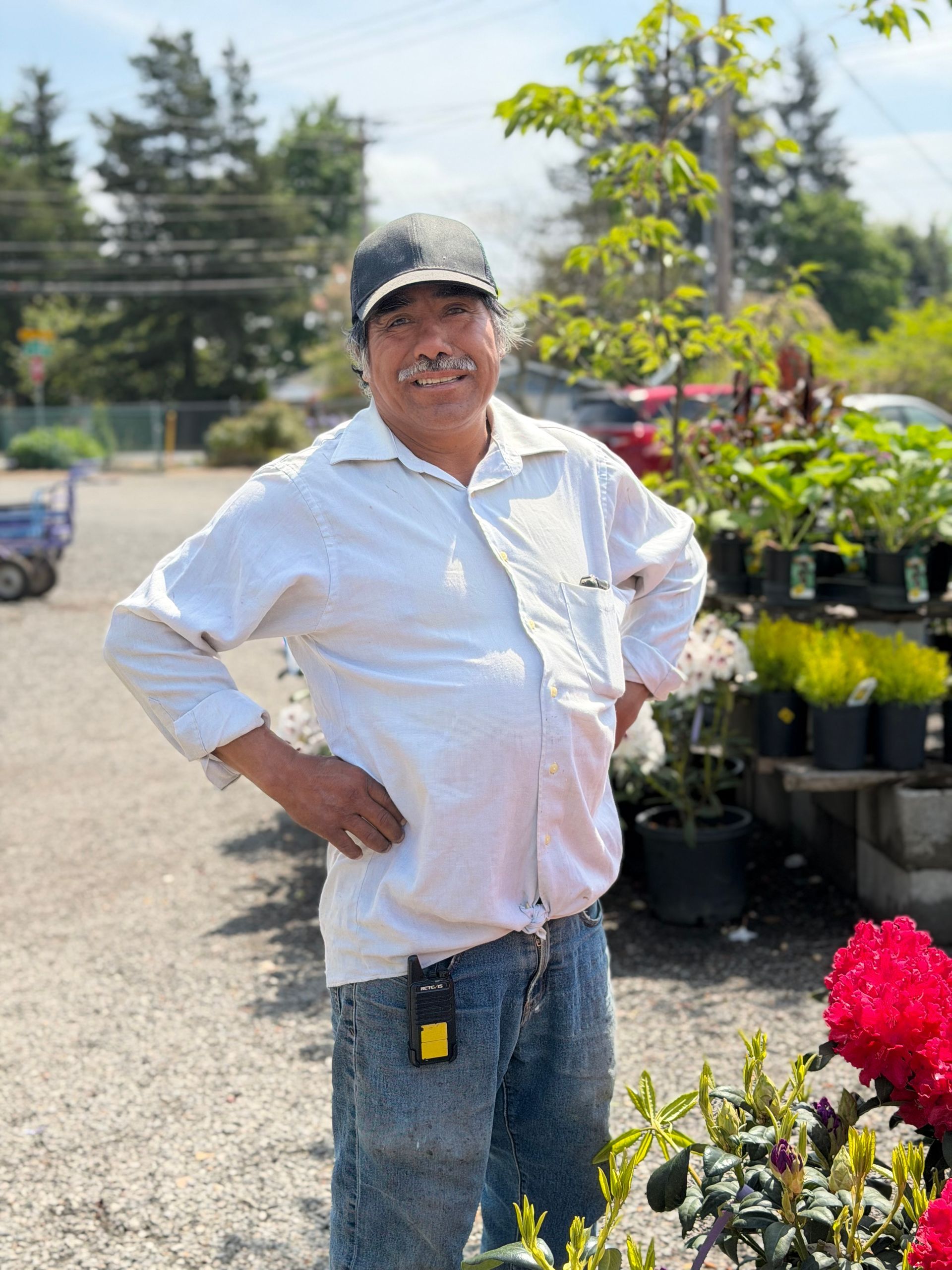 Person standing in a garden center, smiling with hands on hips beside potted flowers and plants.