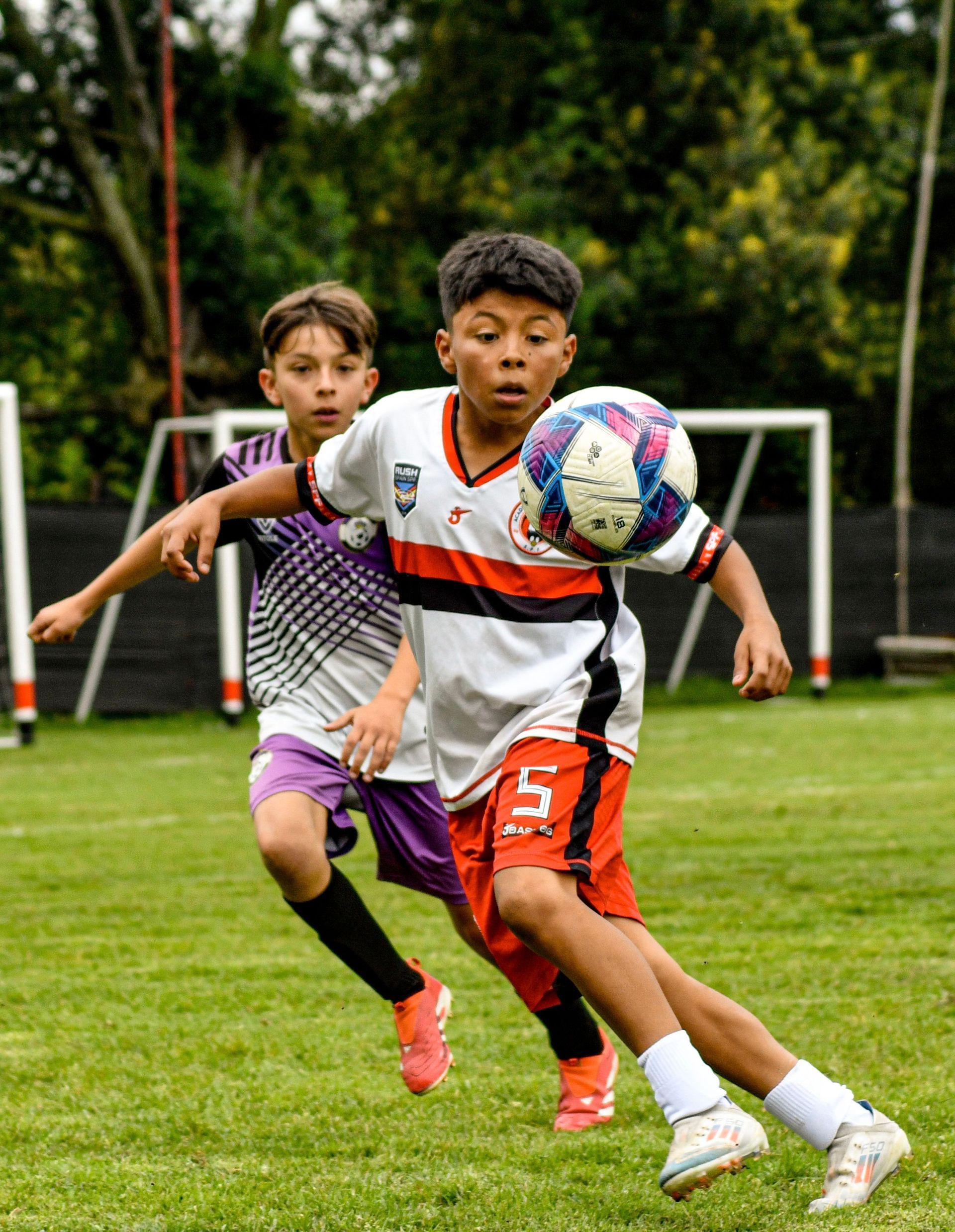 Two soccer players chasing a ball on a grassy field, one in a white jersey in front.