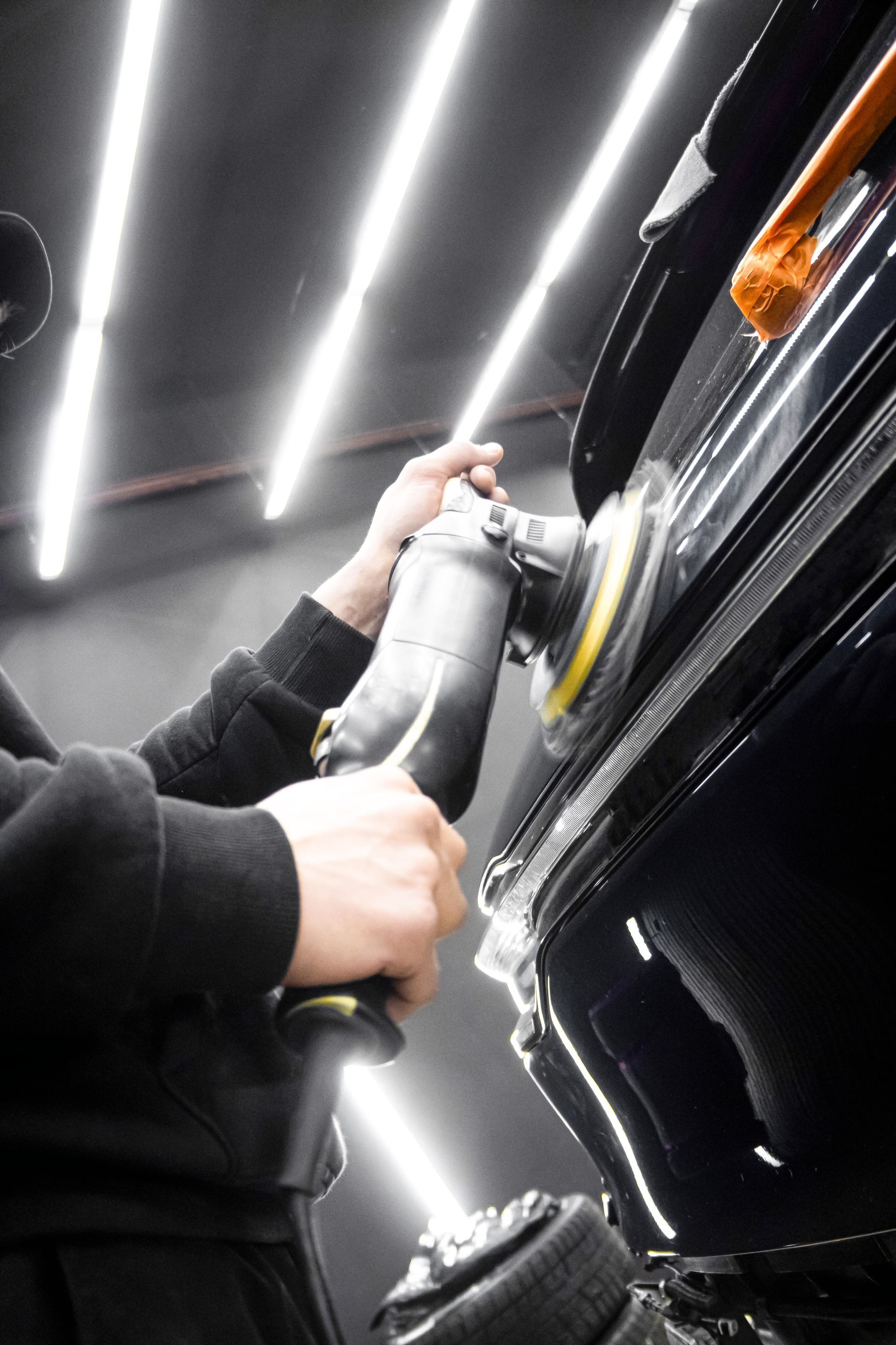 Person polishing a black car with a buffer under bright garage lights