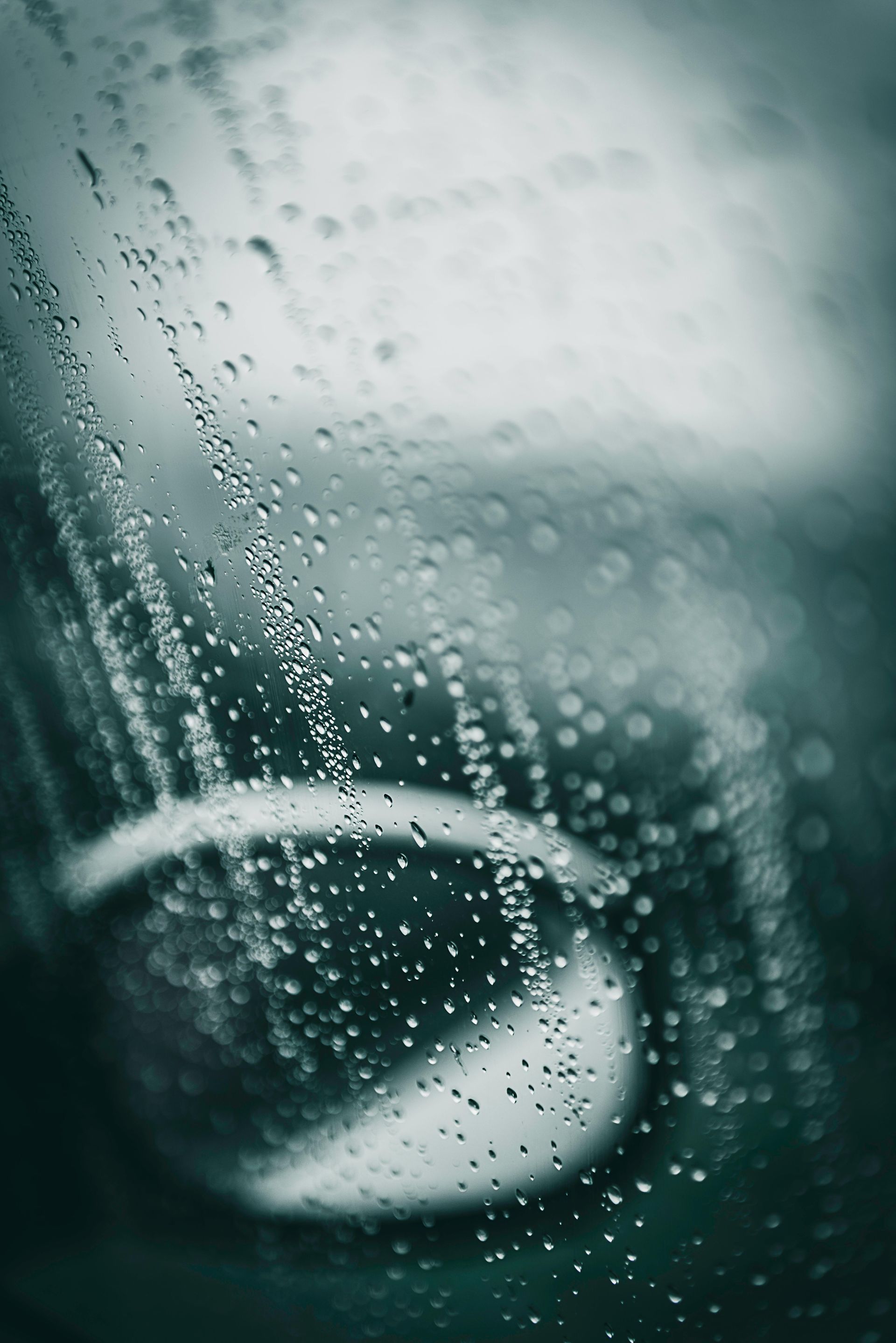Raindrops on glass, blurry view of a car headlight, blue-green tones.