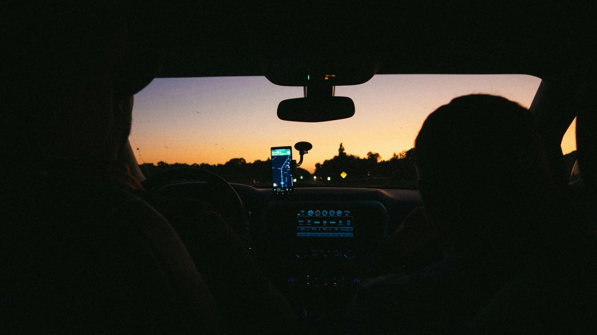Silhouetted figures inside a car during a sunset, viewing a road ahead with a navigation screen visible on the dashboard.