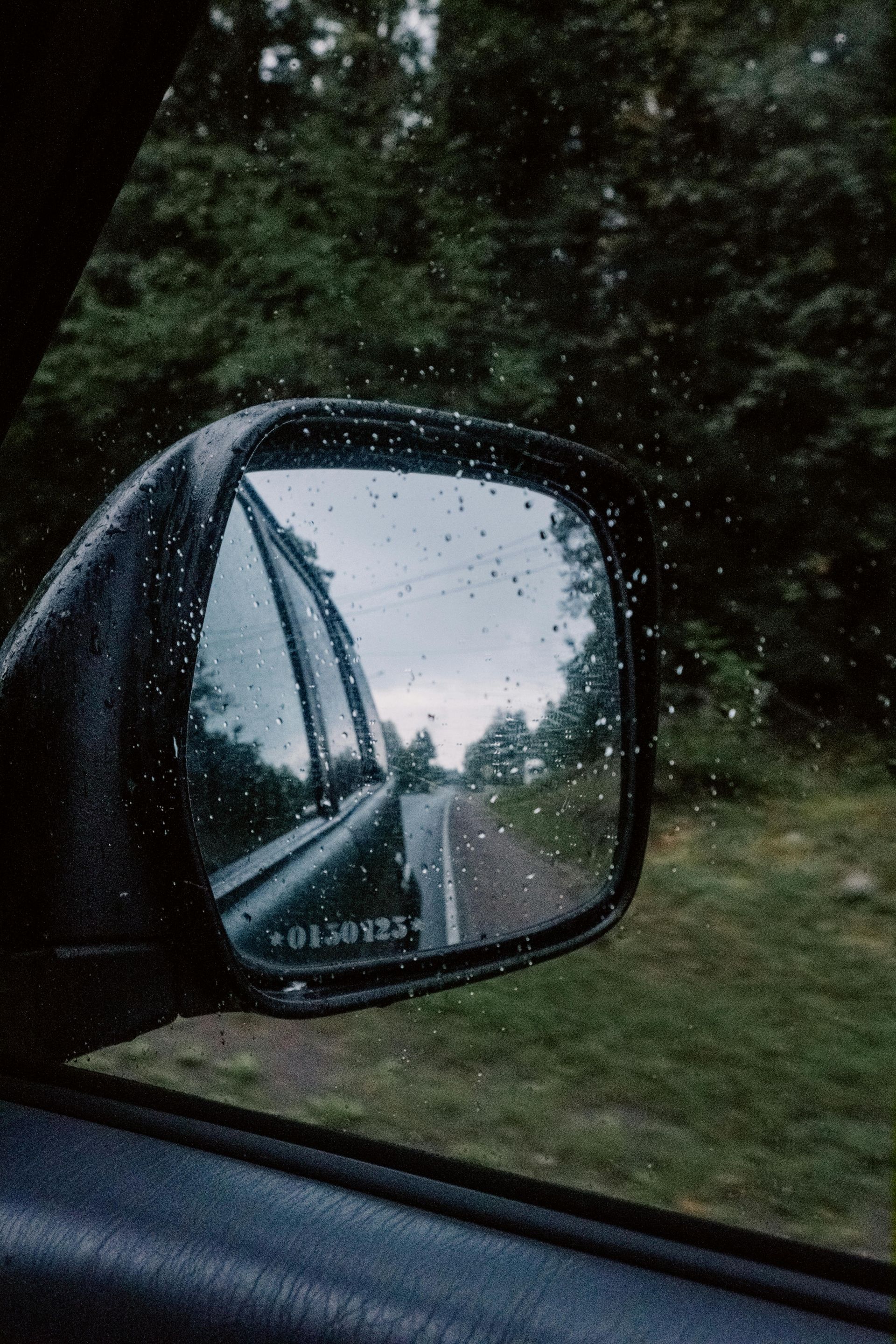 A side-view car mirror reflects a road and forest on a gloomy, rainy day, with droplets covering the glass.
