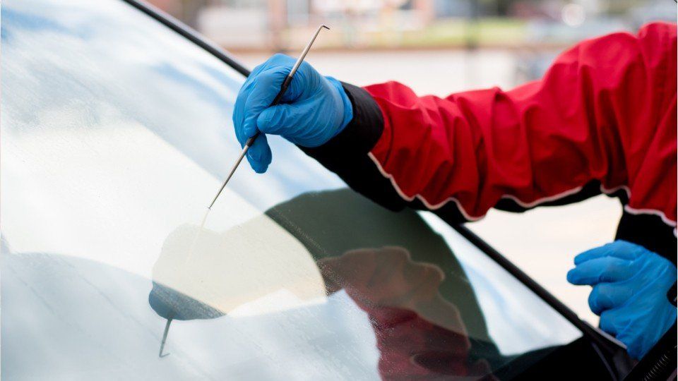Gloved hand with a tool, examining a car windshield. Red jacket, blue gloves, outdoors.