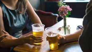 Two people hold plastic cups of beer at a table with a small vase of flowers.
