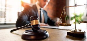 A gavel and balance scales sit on a desk in the foreground, with a person in a suit working in the blurred background.