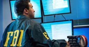 A person wearing an FBI jacket sits at a desk with multiple monitors in a dim, professional office setting.
