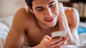 A shirtless person lying in bed, smiling while looking at a smartphone.