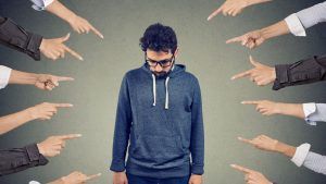 A man wearing glasses and a blue hoodie looks down, surrounded by multiple fingers pointing at him from all sides.
