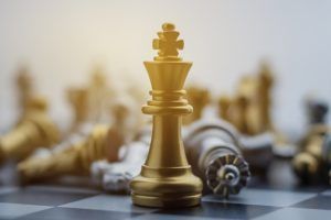 A golden king chess piece standing upright in sharp focus on a chessboard, surrounded by blurred fallen chess pieces.