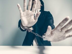 A person in a dark suit with hands raised in a defensive gesture, wearing metal handcuffs connected by a chain.
