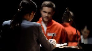 A woman sits across from a person in an orange jumpsuit at a desk, with a lamp illuminating the scene in a dark room.