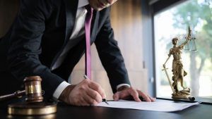 A person in a suit and purple tie signs a document next to a wooden gavel and a gold statue of Lady Justice.
