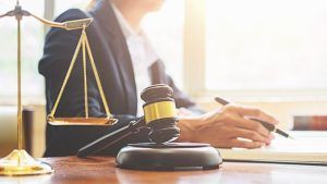A judge’s wooden gavel, a set of golden scales, and a hand writing with a pen on a desk in a law office.
