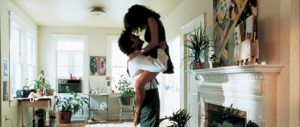 A person lifts another person into their arms in a bright living room with a fireplace, plants, and art.