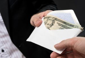 A person in a dark suit hands an open white envelope containing US twenty-dollar bills to another person.