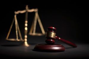 A wooden gavel resting on a sounding block with a golden scale of justice blurred in the background on a black surface.