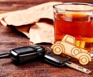 A glass of amber liquid next to a car key, a keychain shaped like a car, and a folded brown paper bag on a wooden surface.