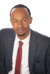 A person in a blue suit, white shirt, and maroon patterned tie looking forward against a plain white background.