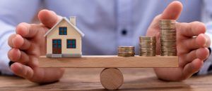 A wooden board balanced on a cylinder, holding a model house on one end and three stacks of coins on the other.