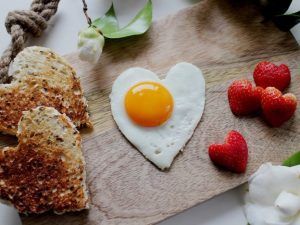 Heart-shaped fried egg, two pieces of toasted heart-shaped bread, and several heart-cut strawberries on a wooden board.