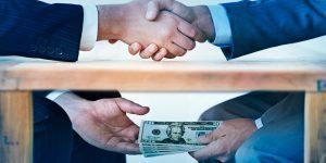 Two people in suits shake hands above a wooden table while one passes a stack of US dollar bills to the other below it.