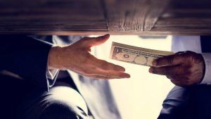 Two people in suits pass a stack of US dollar bills under a table, suggesting a clandestine exchange or bribe.