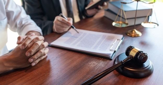 A person sits at a desk with an attorney who points at a document, beside a wooden gavel and scales of justice.