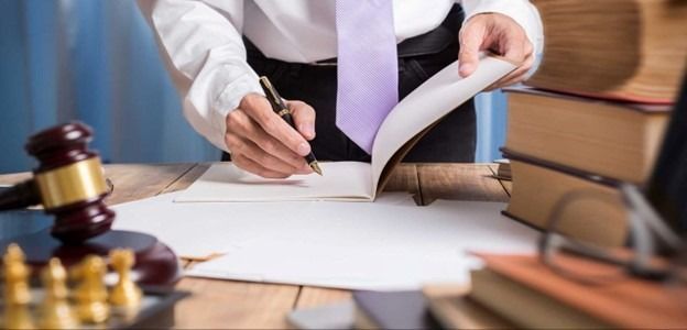 A person in professional attire signs a document at a desk with a gavel, a chess set, and stacked law books nearby.