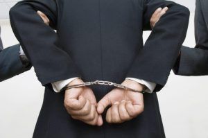 A person in a dark suit being held by their arms while wearing metal handcuffs behind their back against a plain background.