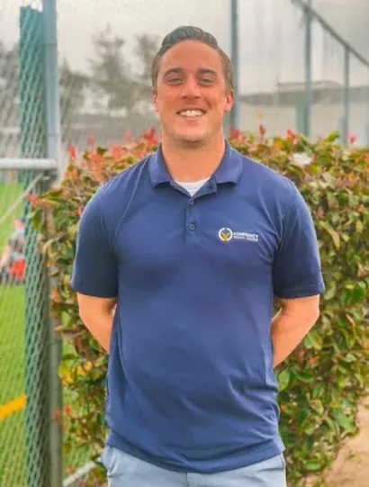 Smiling man in a blue polo shirt standing outdoors in front of a hedge and chain-link fence