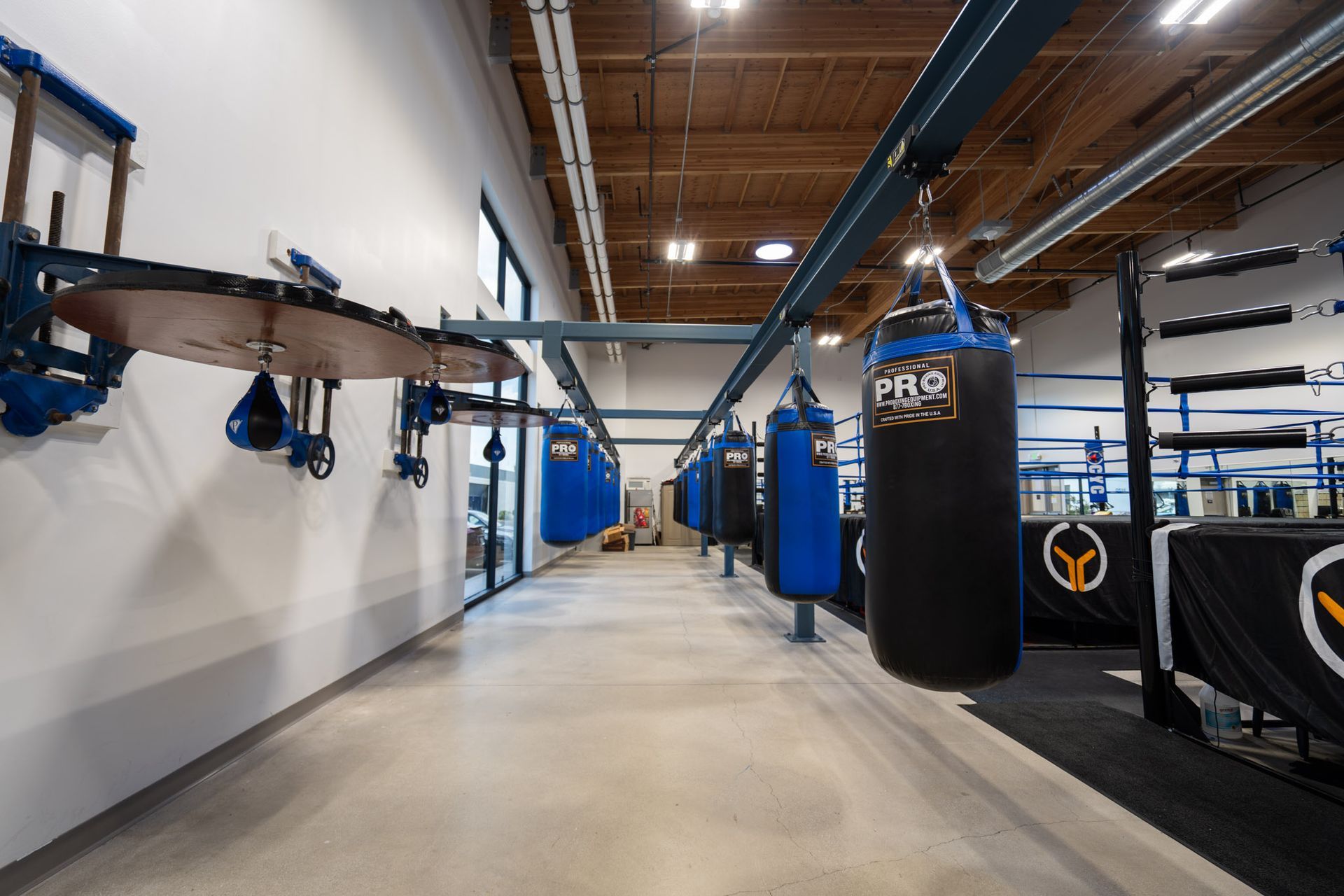 Wide indoor gym with heavy bags, suspension rigs, and blue boxing equipment along a long hallway