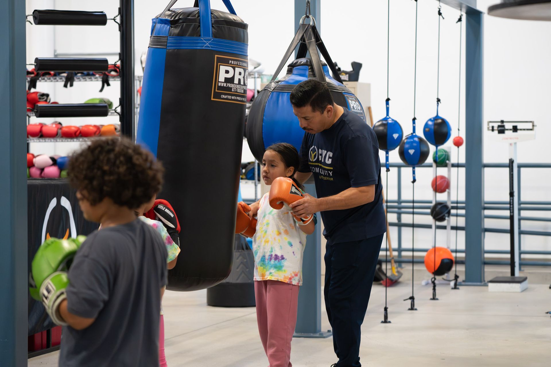 Gym scene with a coach helping a child near a hanging punching bag and boxing equipment.